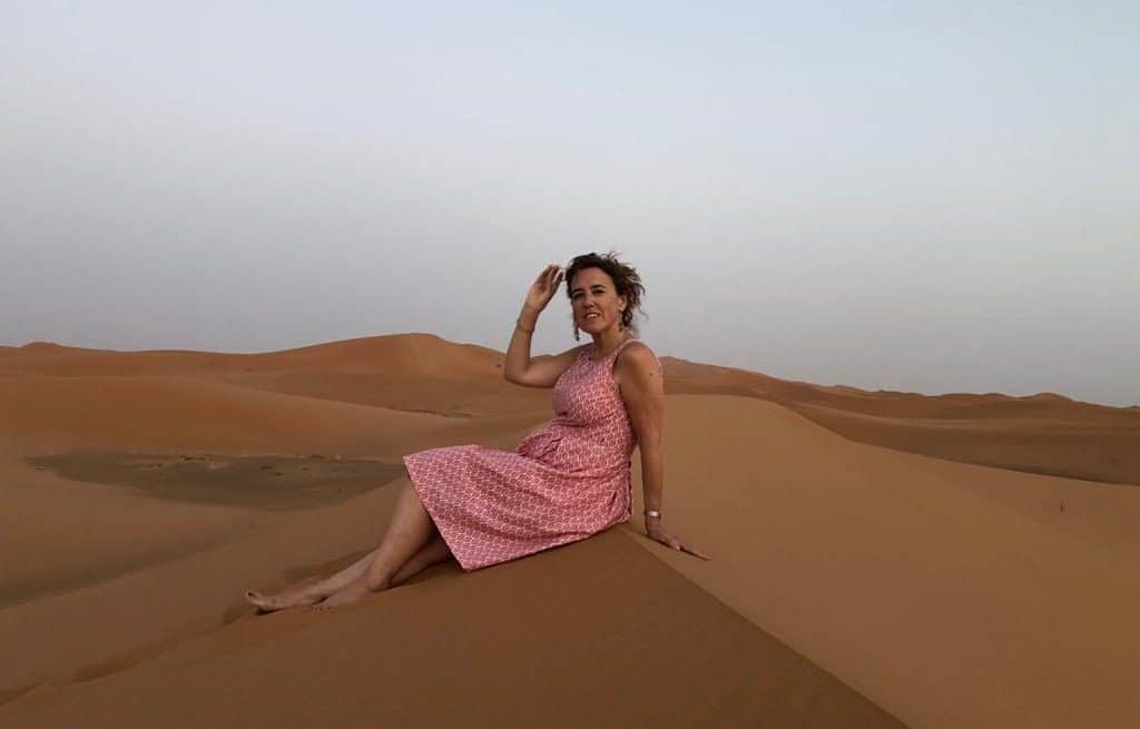 Una persona con un vestido rosa se sienta en una duna de arena en un paisaje desértico bajo un cielo despejado.