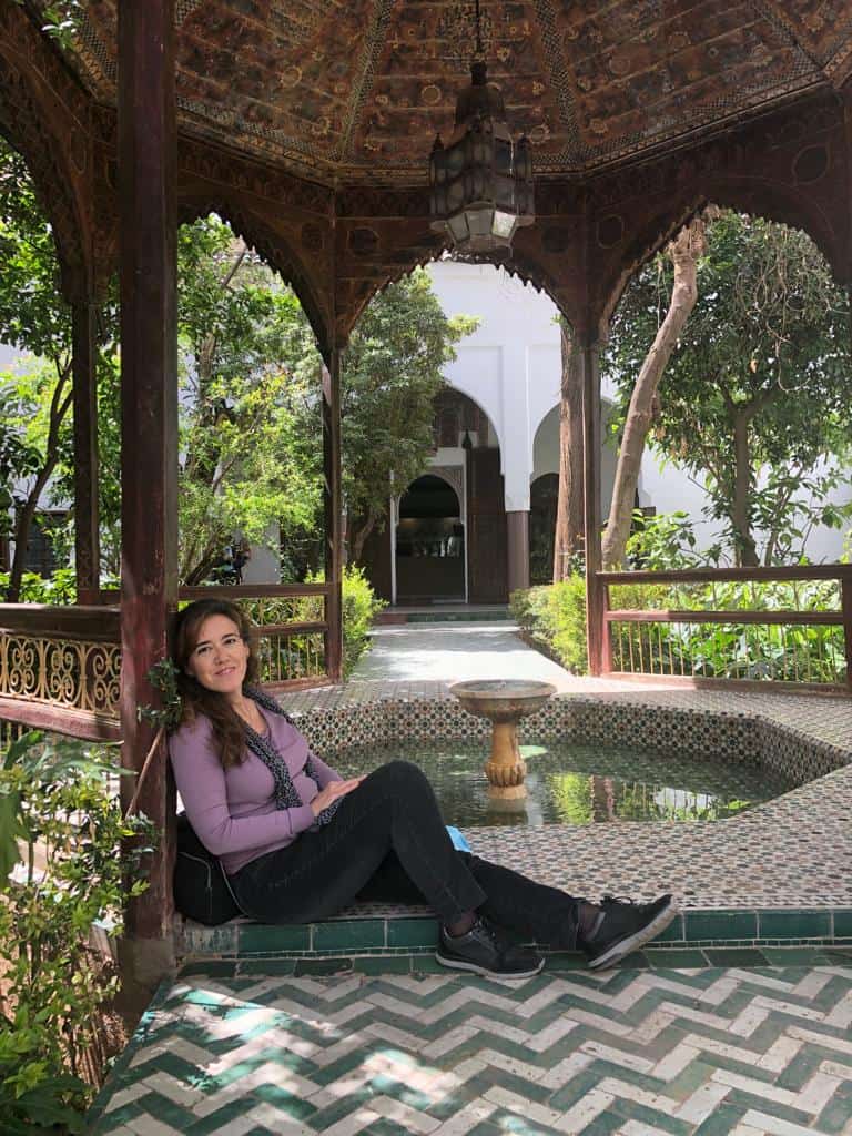 Una persona sentada en una cornisa de baldosas en un mirador ornamentado, rodeado de vegetación y una fuente, con un patio abierto visible al fondo.