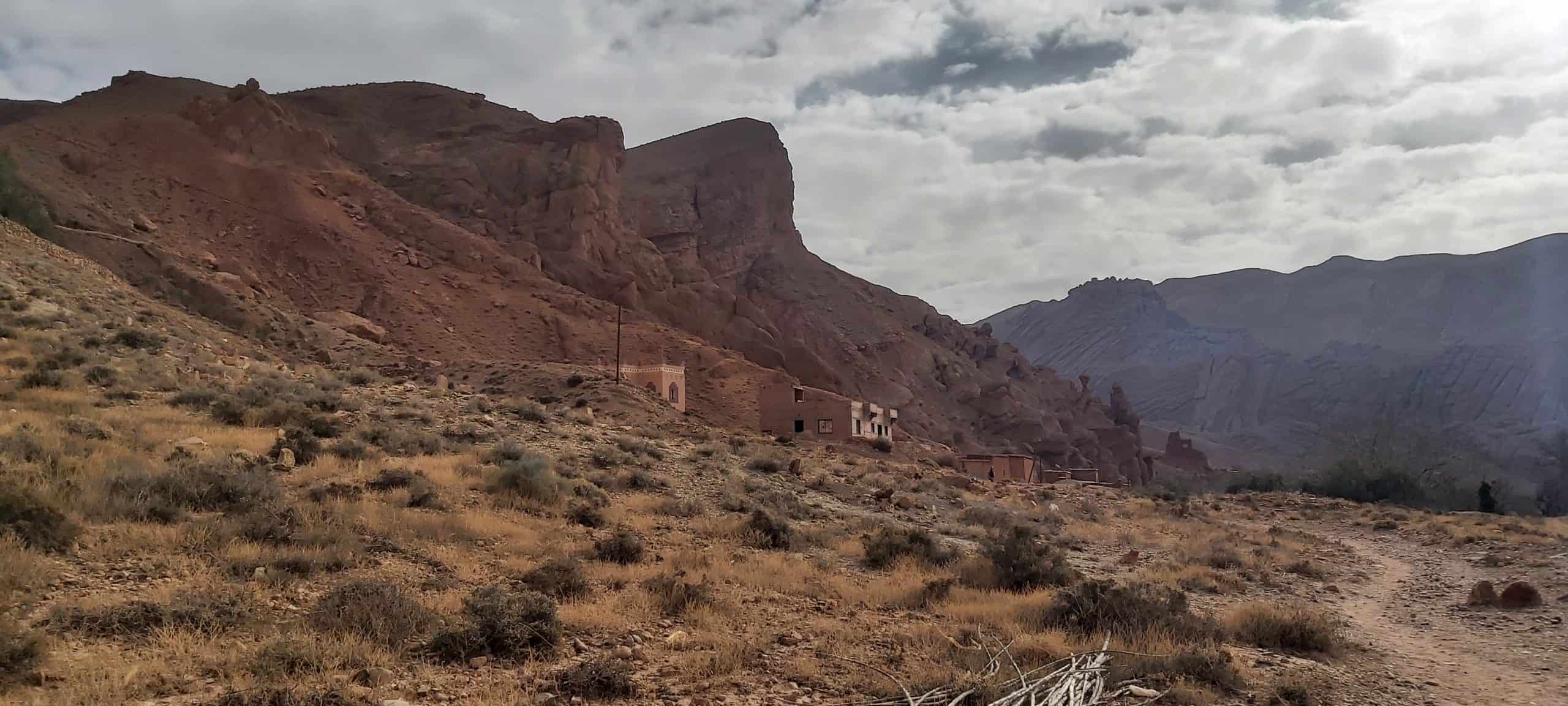 Paisaje árido con arbustos dispersos, colinas rocosas y varios edificios pequeños en la base de una montaña empinada bajo un cielo nublado.