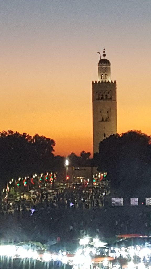 Una torre alta se alza contra un cielo anaranjado del atardecer, con árboles, luces y una multitud de personas y puestos de mercado en primer plano.