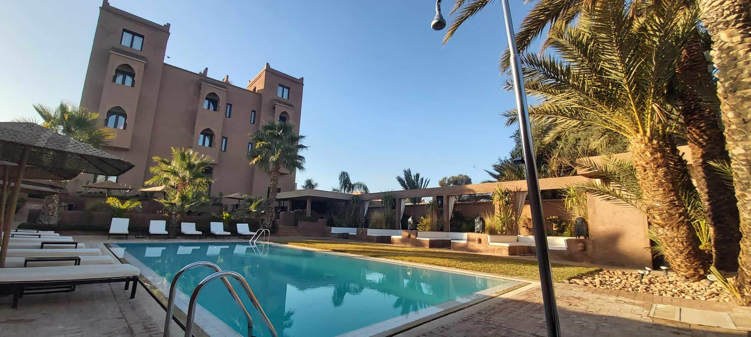 Una piscina rectangular al aire libre rodeada de tumbonas y palmeras, con un edificio de varios pisos y una zona de estar con sombra al fondo bajo un cielo despejado.