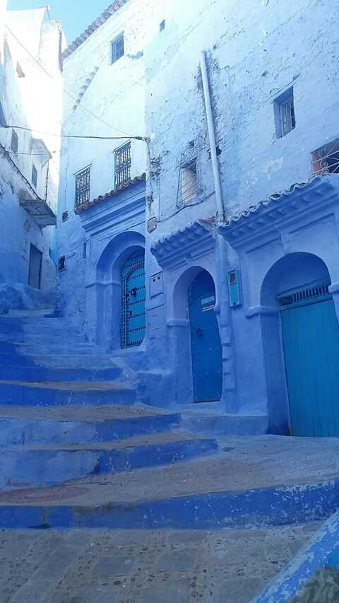 Carreró estret amb escales, portes i parets pintades de blau en una zona residencial, probablement a Chefchaouen, Marroc, sota la llum del dia.