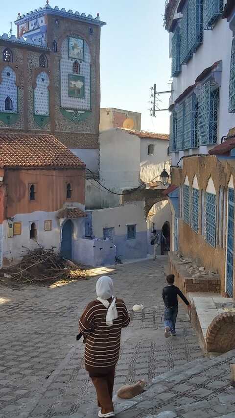 Una dona i una nena caminen per un carrer empedrat amb edificis blaus i blancs en un barri històric assolellat.