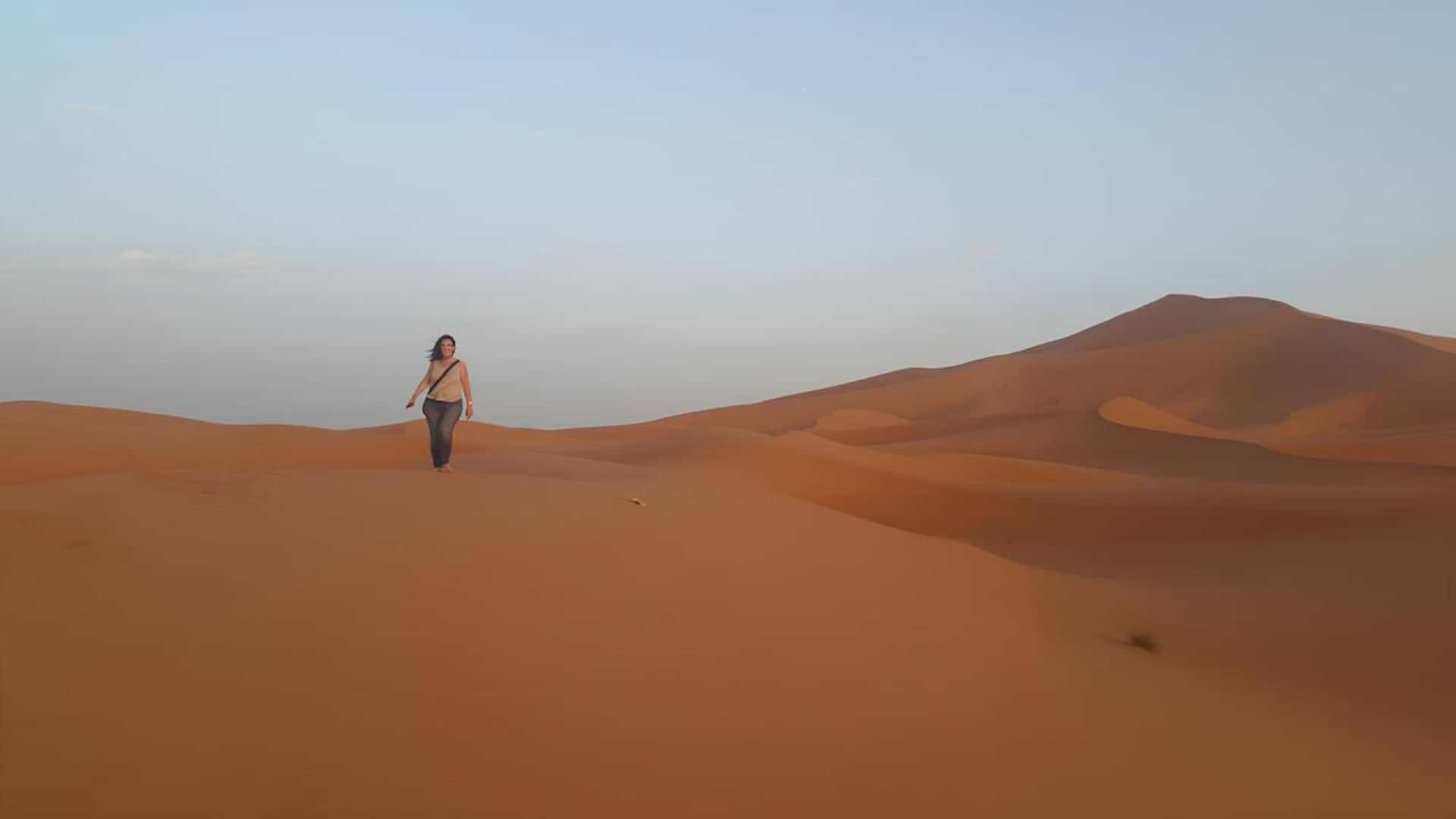 Una persona camina sola per dunes de sorra taronja sota un cel clar, amb suaus turons de sorra al fons.
