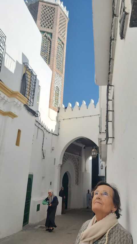 Una dona mira cap amunt en un carreró blanc i estret amb portes arquejades i un alt minaret de mesquita al fons sota un cel blau clar.