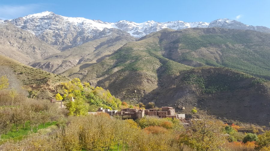 Un petit poble amb edificis marrons es troba a la base d'una serralada verda, amb cims nevats visibles al fons sota un cel blau clar.