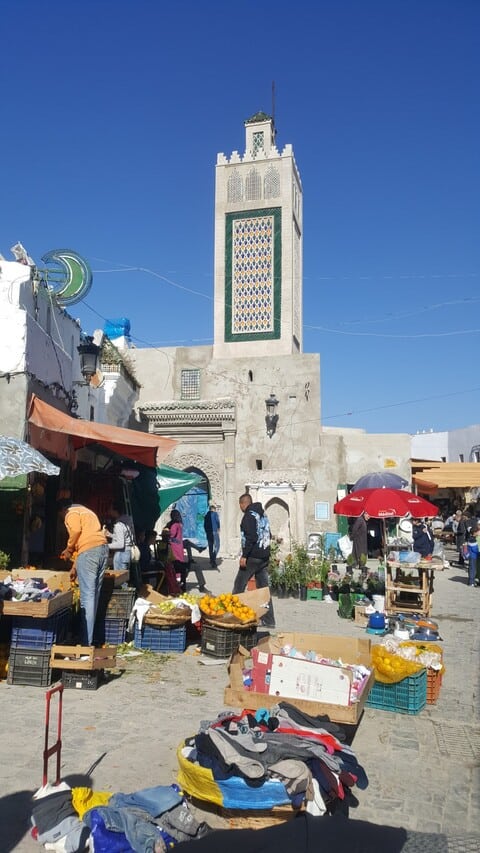 Un mercat a l'aire lliure amb parades de productes frescos i roba davant d'una alta torre de mesquita sota un cel blau clar. La gent compra i passeja.