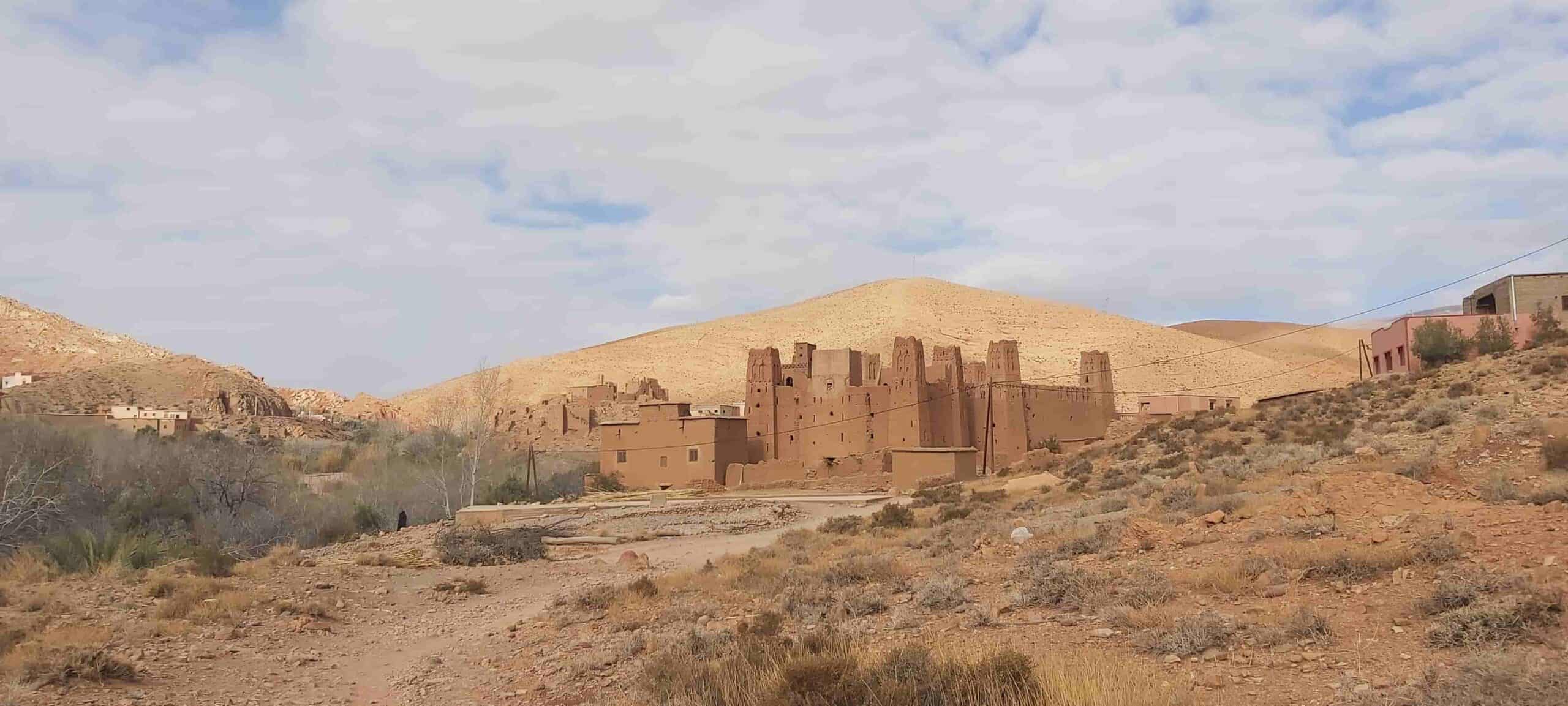 Un antic poble de tova amb edificis alts i marrons s'alça en un paisatge sec i rocós sota un cel parcialment ennuvolat.